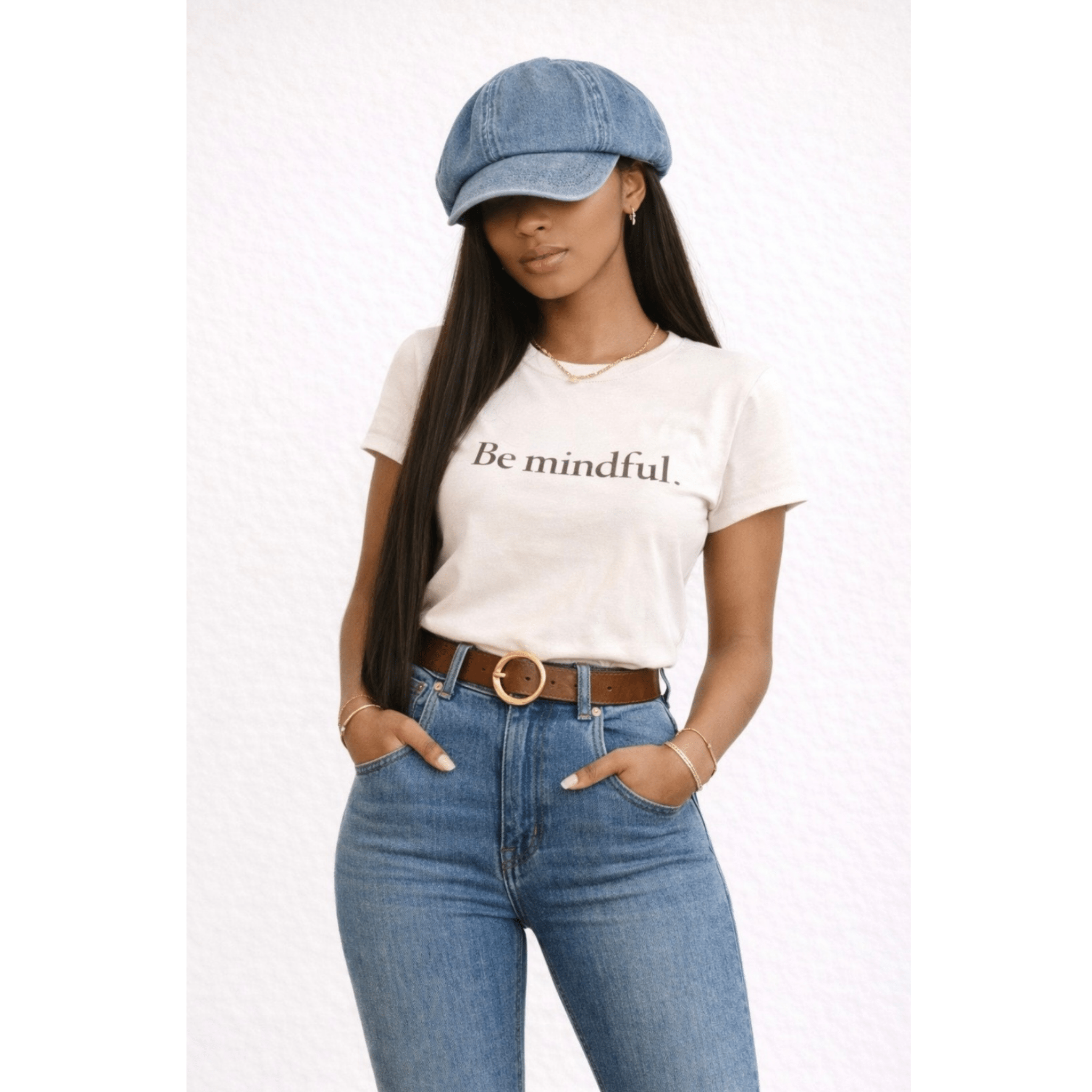Woman wearing a beige t-shirt with 'Be mindful' text, blue jeans, and a denim cap on a white background