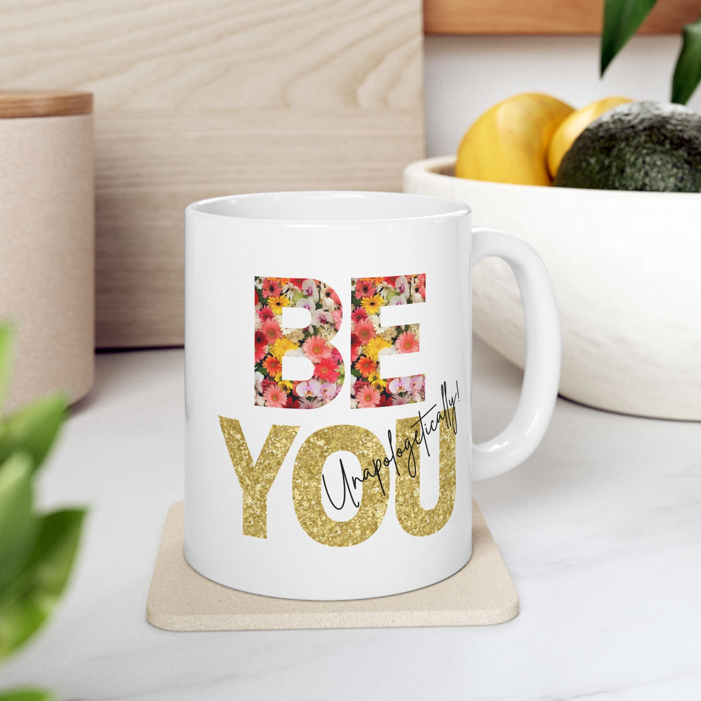 White ceramic mug with 'BE YOU Unapologetically' inspirational text on a coaster, surrounded by plants and a bowl of fruit.