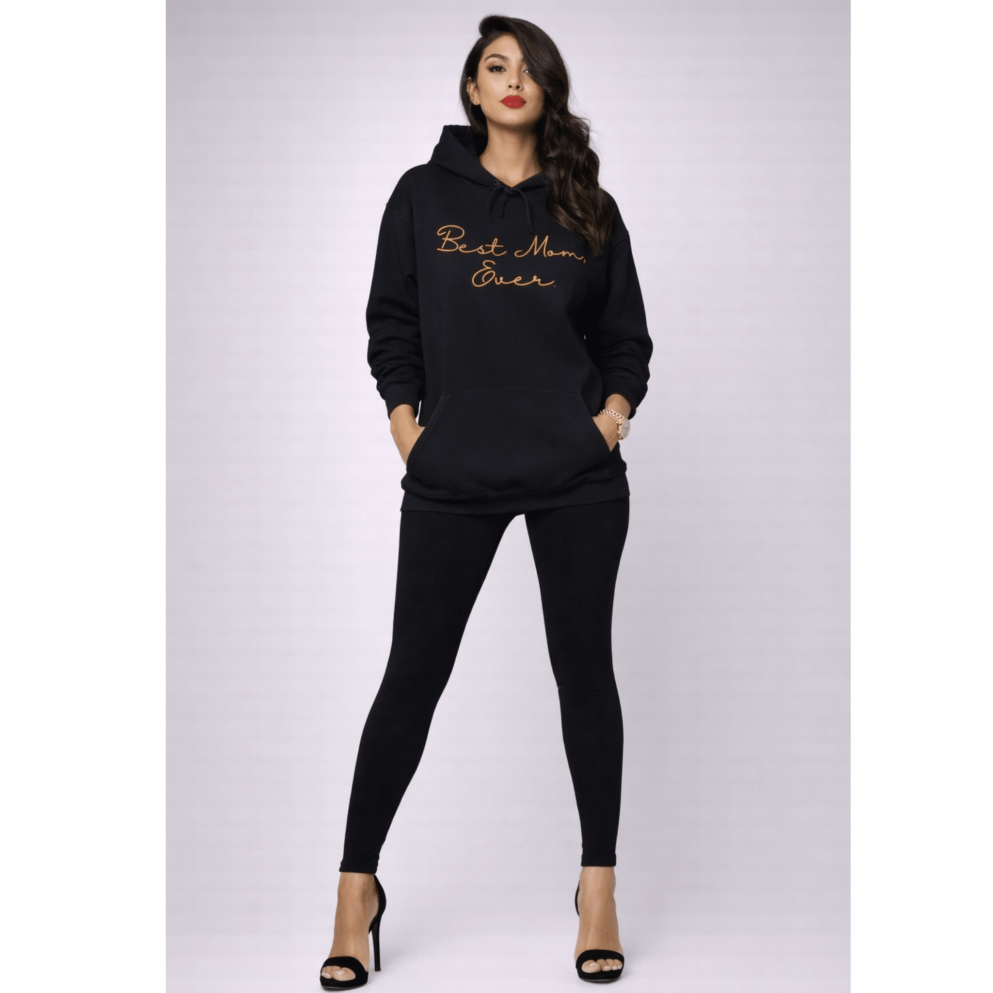 Woman wearing a black hoodie with 'Best Mom Ever' text with black leggings on a white background