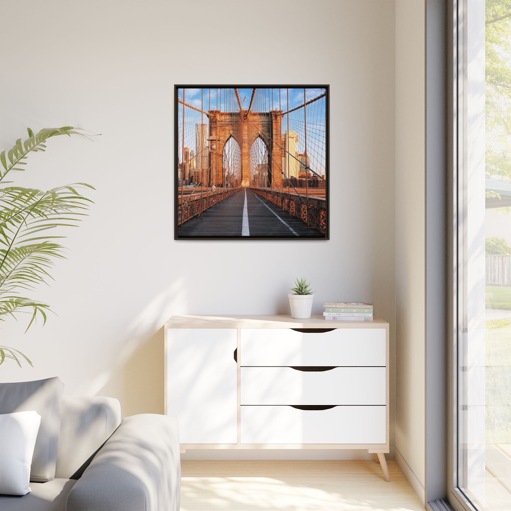 Framed picture of Brooklyn bridge on a wall above a white cabinet with a plant and books.