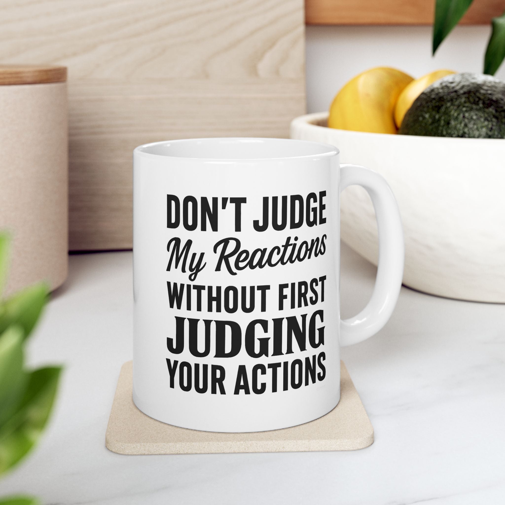 White ceramic mug with bold quote about reactions and actions in black text on a coaster with fruit in the background.