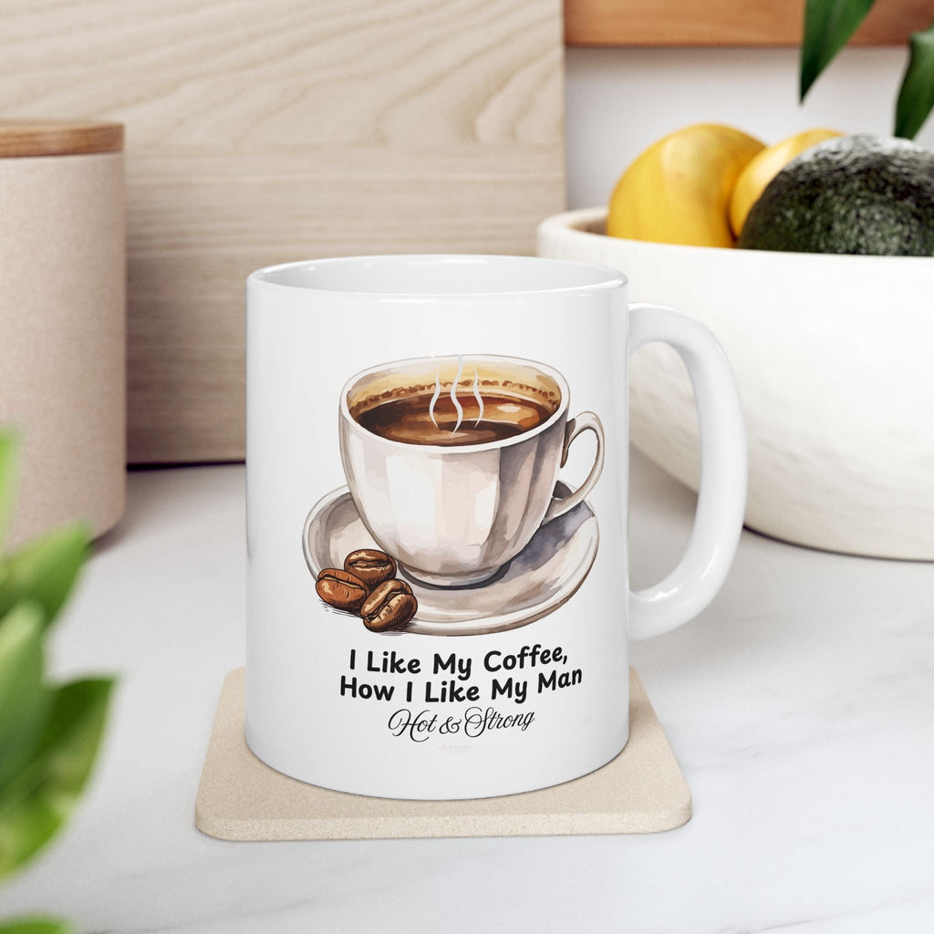 White mug with coffee cup design and text on a coaster, placed on a kitchen counter surface near bowl of fruit in background.