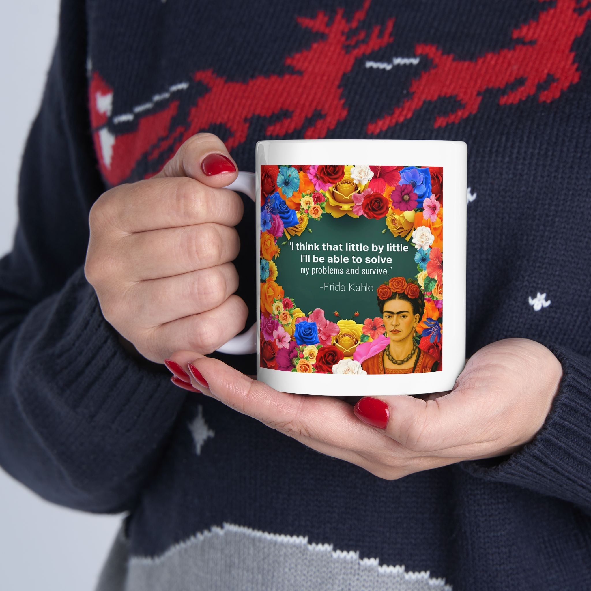 Person holding a floral mug with Frida Kahlo design and quote, wearing a sweater with reindeer pattern.