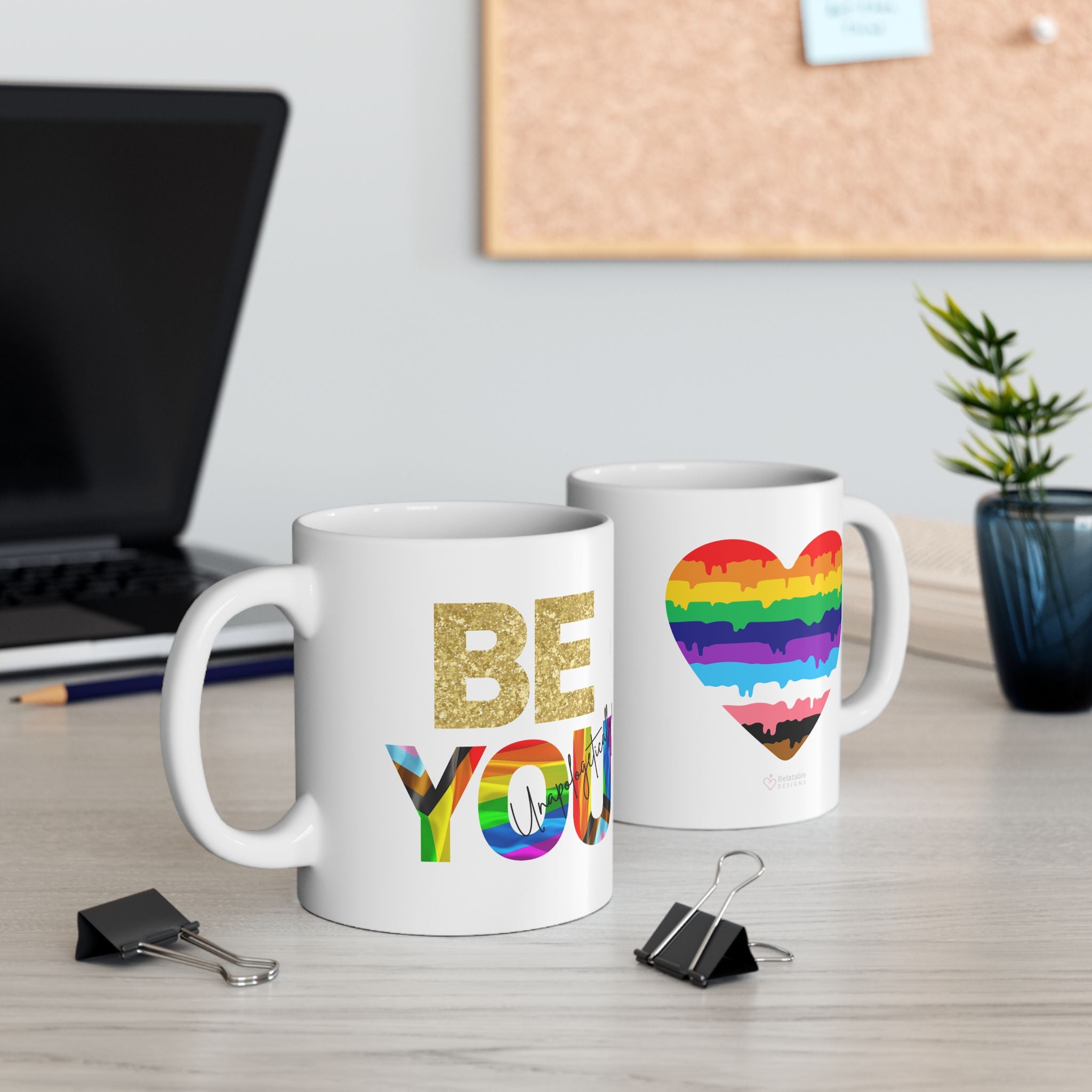 Two mugs on a desk with one displaying 'BE YOU' in rainbow letters and the other with a rainbow heart design.