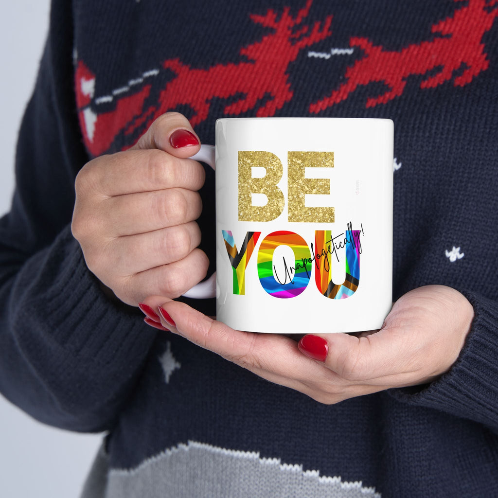 Person holding a mug with 'BE YOU' text in colorful LGBTQIA+ letters, wearing a dark sweater with red deer designs.