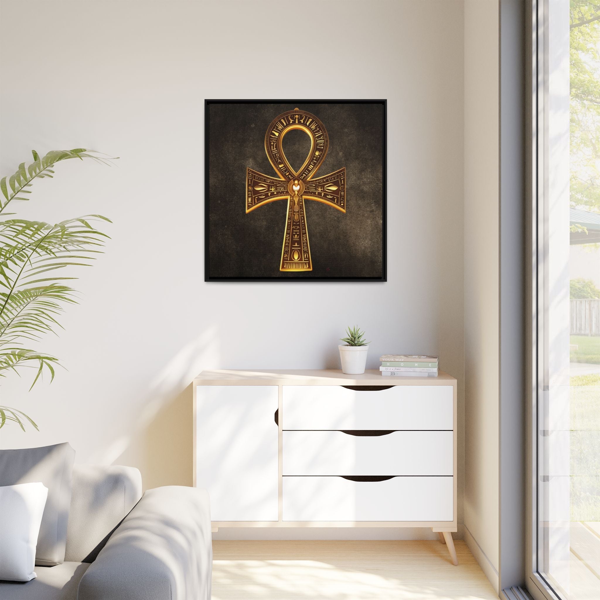 Decorative gold ankh artwork on a wall above a white cabinet with a plant and window in the background.