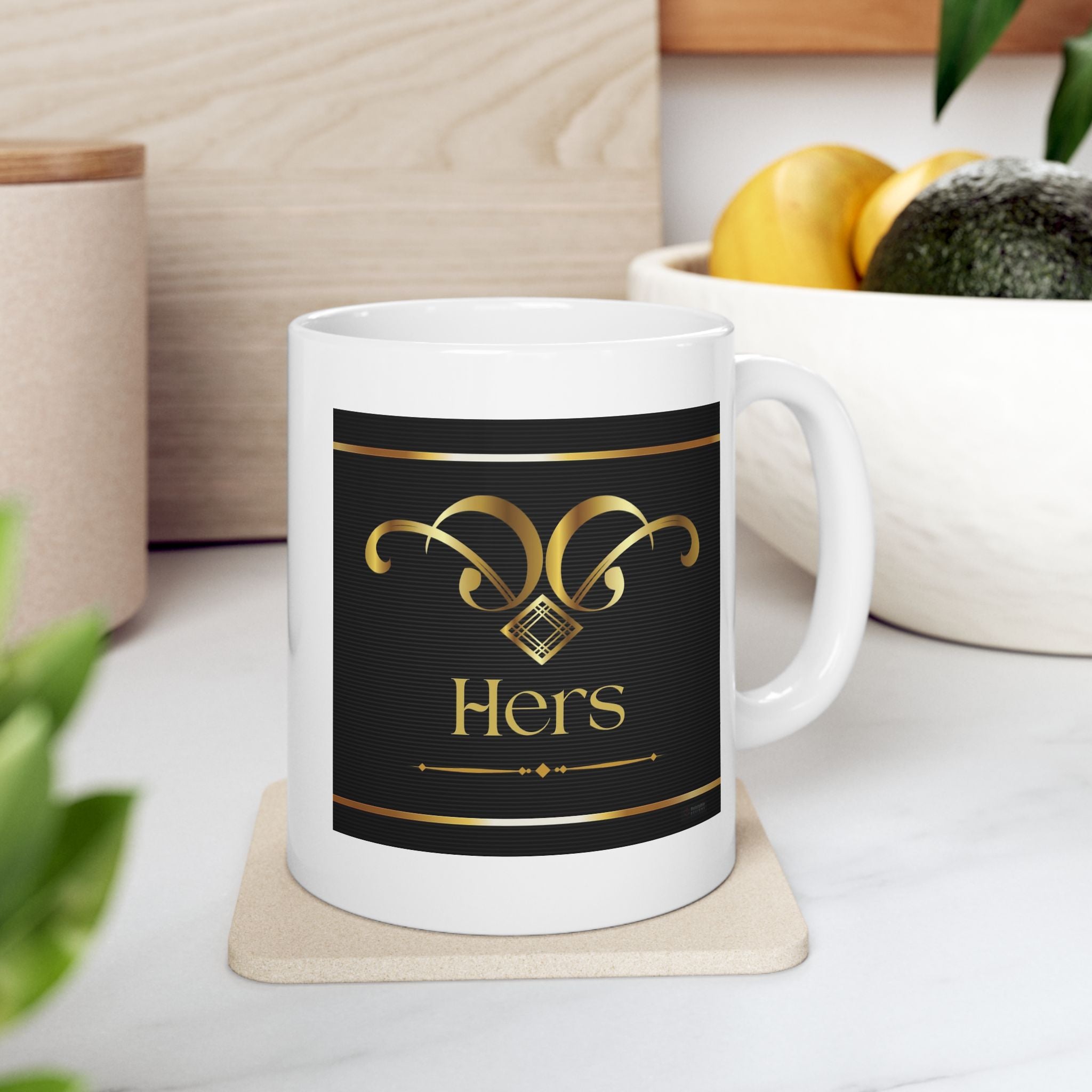 White ceramic 'HERS' mug with gold and black design on a coaster, surrounded by decorative items.