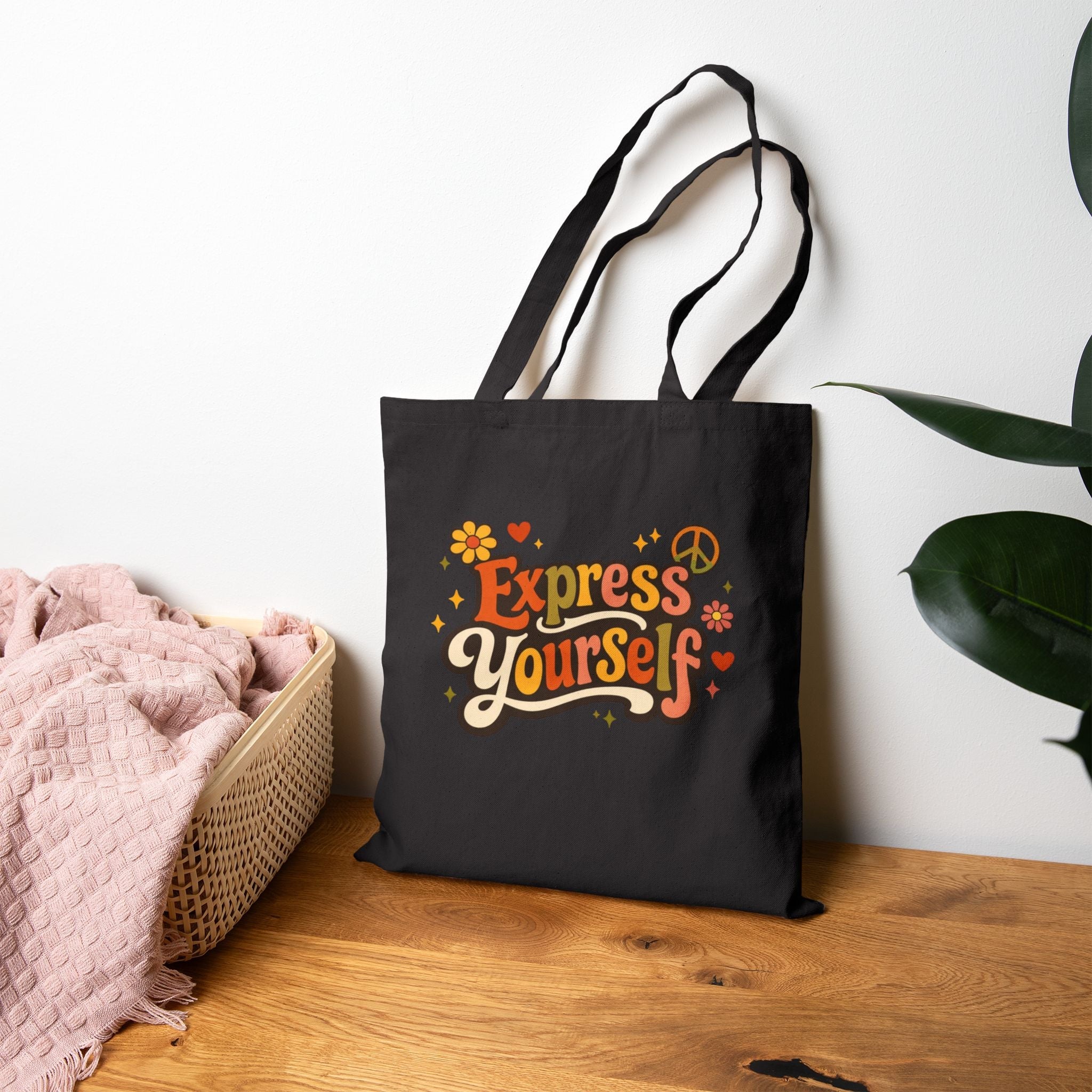 Black tote bag with 'Express Yourself' text on a wooden surface with a plant in the background