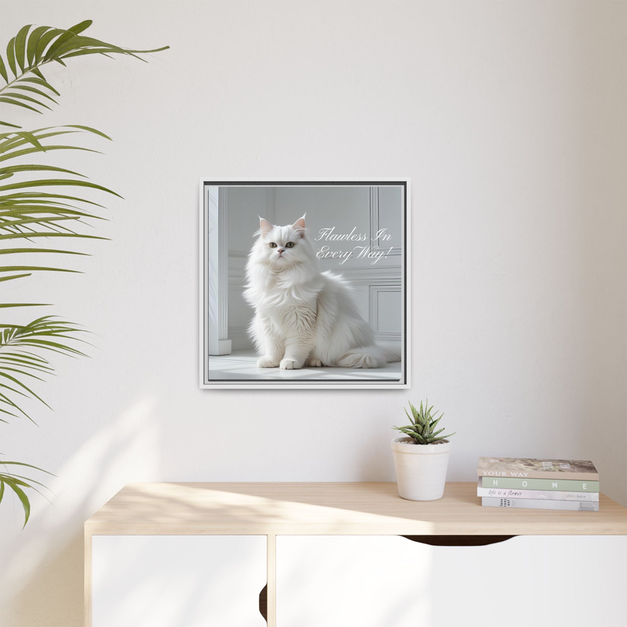 Framed picture of a white cat with text 'Flawless In Every Way' on a wall above a desk with a plant and books.