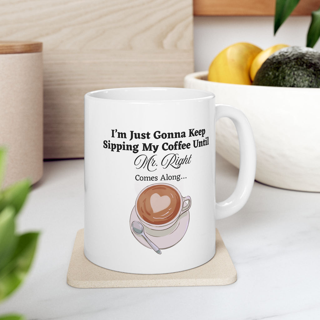 White mug with coffee graphic and text on a coaster placed on a kitchen counter with bowl of fruits in background. 