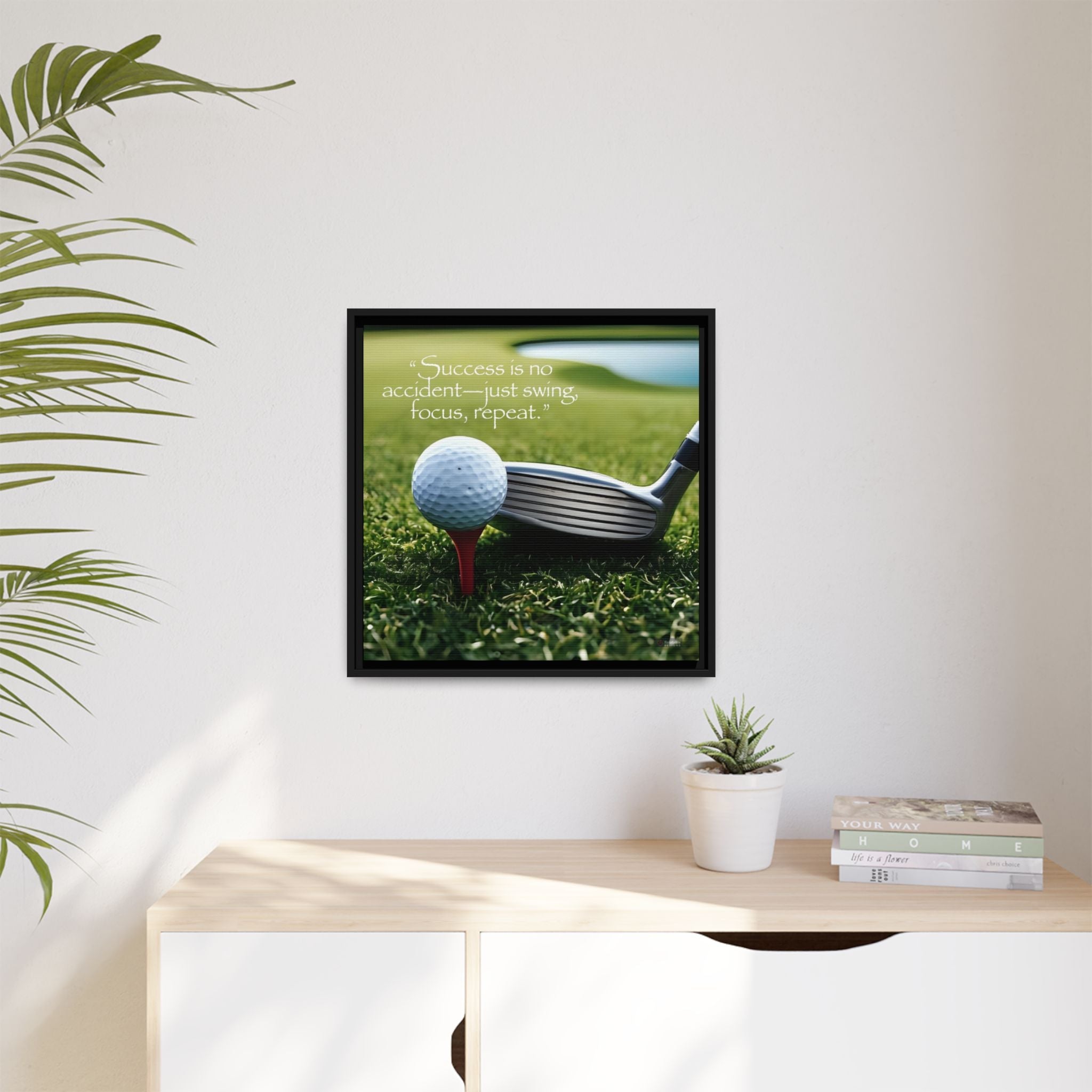 Framed image of a golf ball and club on grass with a motivational quote, placed on a desk with a plant and books.