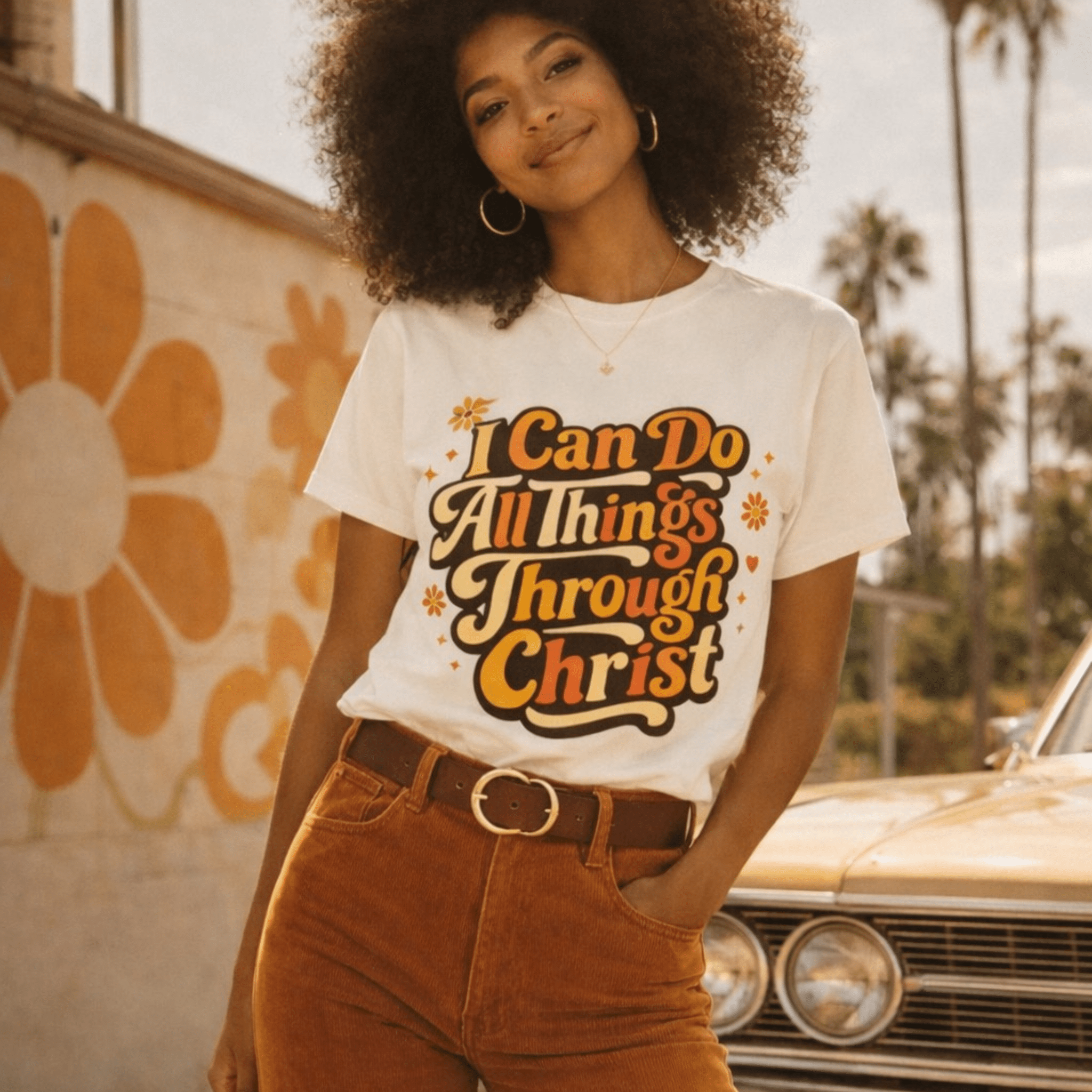 Woman wearing a beige t-shirt with a motivational quote, standing in front of a vintage car and wall with a large flower design.