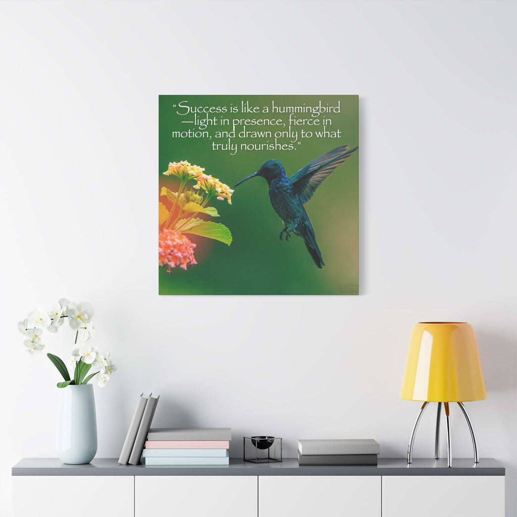 Wall art with a hummingbird and inspirational quote on a green background, displayed above a table with books and a lamp.