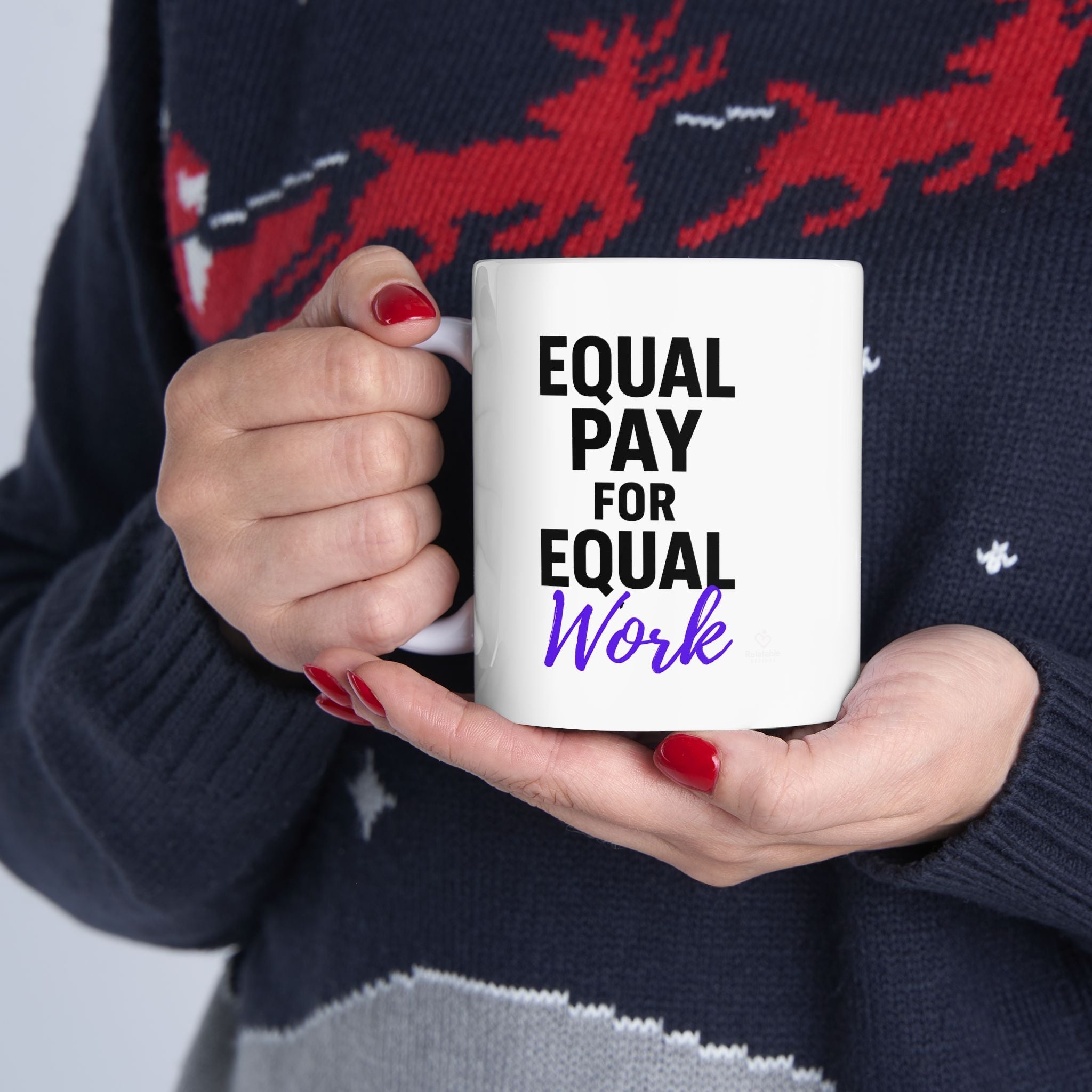 Person holding a mug with 'Equal Pay for Equal Work' text, wearing a dark sweater with red deer designs.