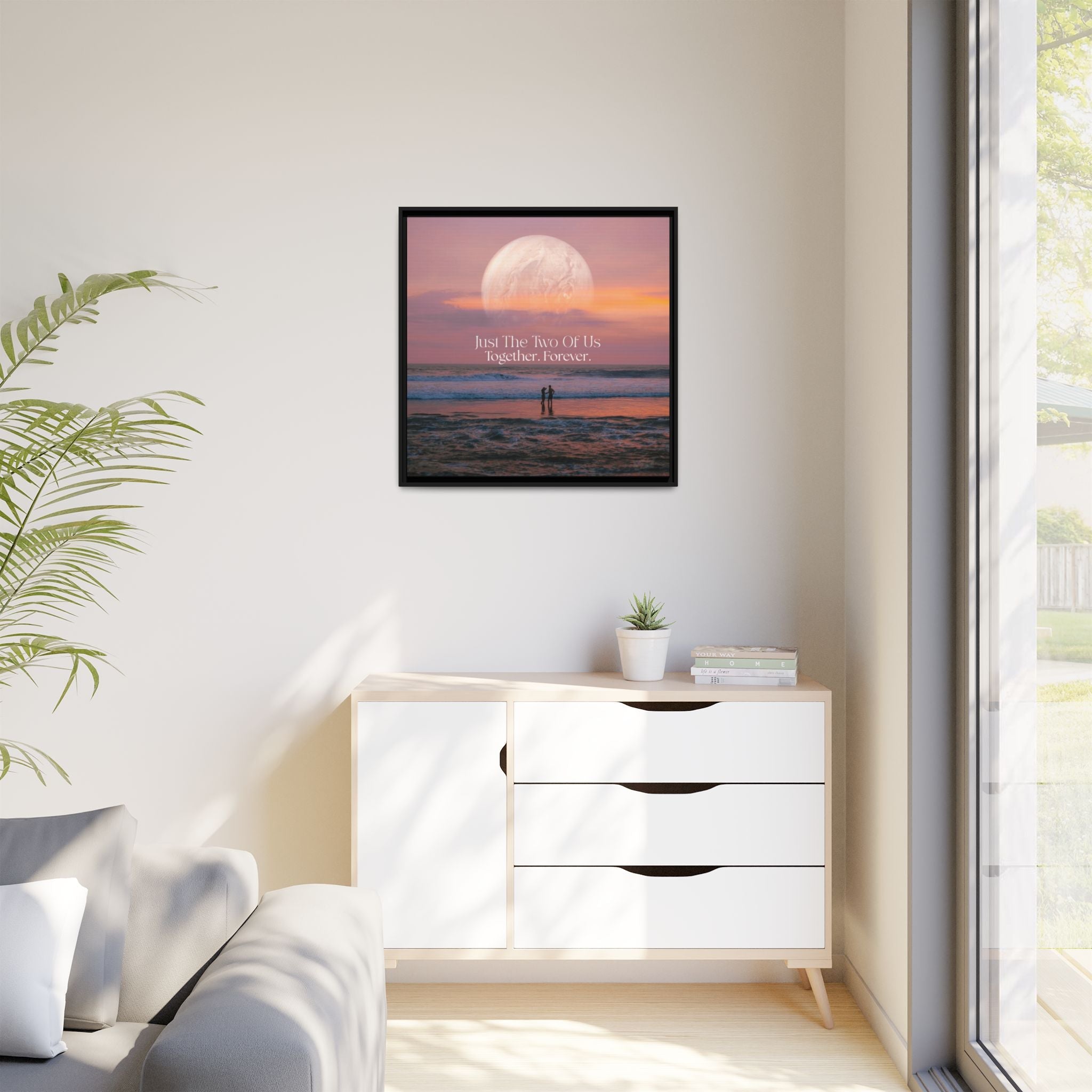 Framed romantic artwork on a wall above a white dresser with a plant and window in the background.