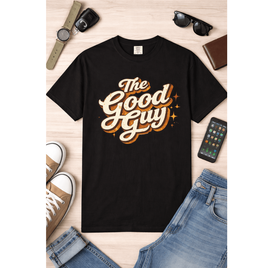 Black t-shirt with 'The Good Guy' text on a wooden surface with casual clothing items for the perfect outfit.