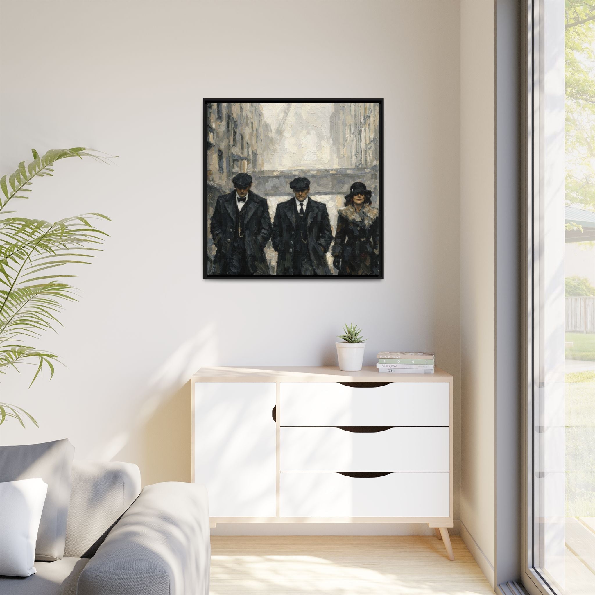 Framed artwork of three people on a wall above a white cabinet with a plant and books.